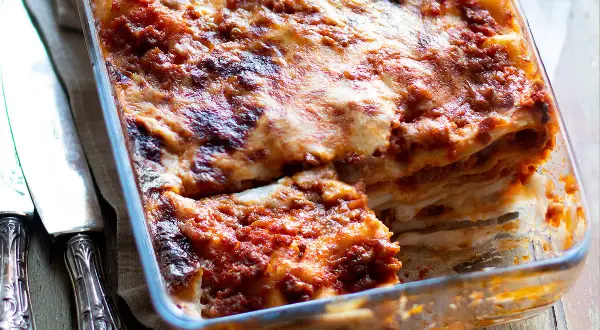 Lasagne alla bolognese appena sfornate, dorate e filanti, in una teglia di vetro. Accanto, due coltelli d'argento.
