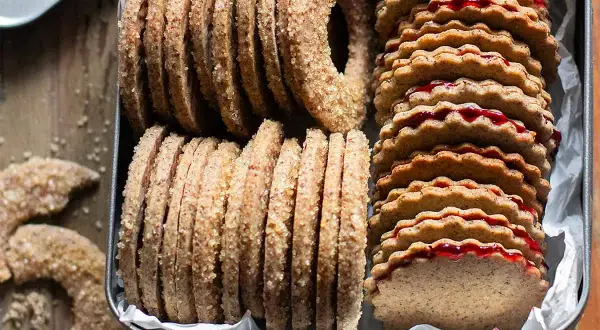 Biscotti grano saraceno ripieni di marmellata, impilati in una scatola di latta. Alcuni biscotti a forma di ciambella e a mezzaluna sono sparsi sul tavolo.
