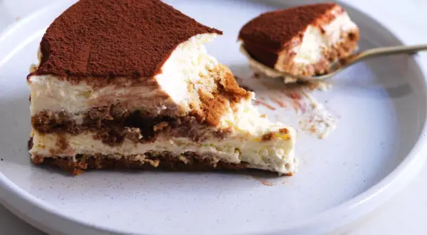 Una fetta di tiramisù ricetta originale, con strati di savoiardi, crema al mascarpone e cacao in polvere. Un cucchiaino ne ha già assaggiato un pezzetto.