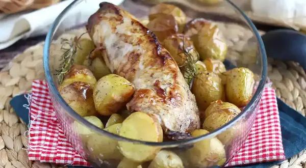 Un succulento filetto di maiale al forno, dorato e profumato, circondato da patate novelle arrosto e rametti di rosmarino in una ciotola di vetro.