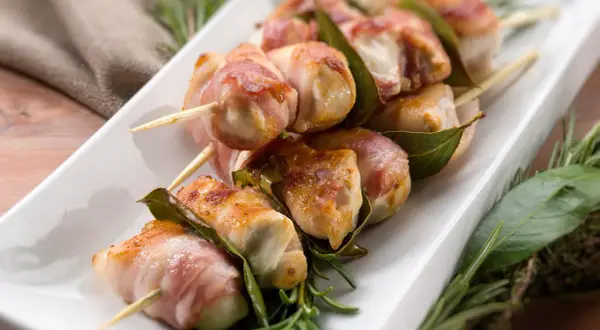 Involtini di lonza avvolti in pancetta, infilzati su spiedini con foglie di alloro e rosmarino.