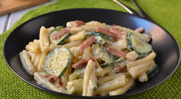 Piatto di pasta cremosa con zucchine a rondelle e cubetti di speck croccante, guarnito con pepe nero.
