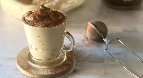Deliziosa crema al caffè, spolverata di cacao, servita in una tazza di vetro con un colino per caffè accanto.