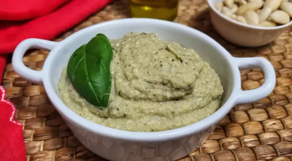 Pesto di melanzane cremoso in una ciotola bianca, guarnito con una foglia di basilico. Sullo sfondo, mandorle e olio d'oliva.