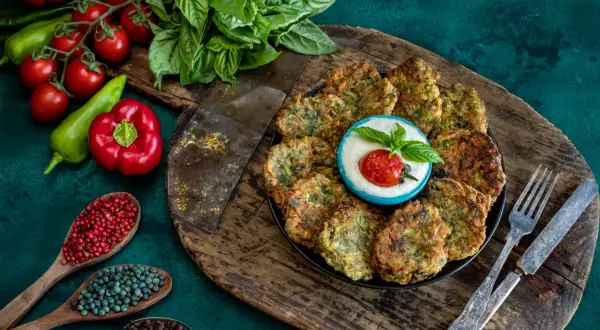 Piatto di polpette di zucchine dorate, servite con una ciotolina di salsa bianca, pomodorino e basilico. Ingredienti freschi e spezie completano la scena.
