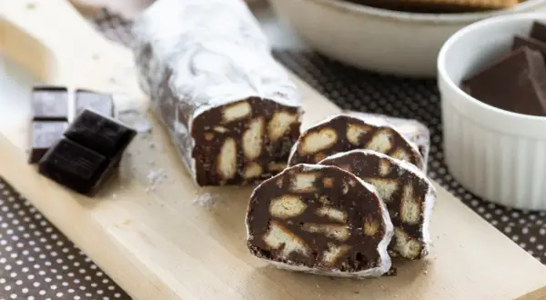 Un delizioso salame al cioccolato, cosparso di zucchero a velo, con biscotti e cioccolato fondente a lato.