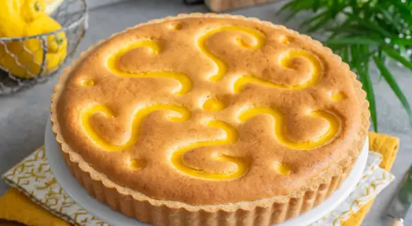 Torta con crema pasticcera, decorata con un motivo a spirale giallo brillante. Sullo sfondo, limoni freschi e foglie verdi.