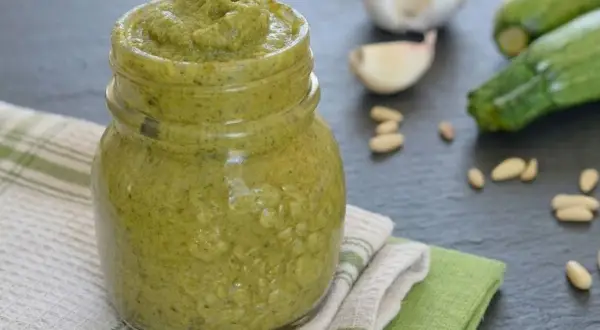 Un vasetto pieno di cremoso pesto di zucchine, con ingredienti freschi come zucchine, aglio e pinoli sullo sfondo.