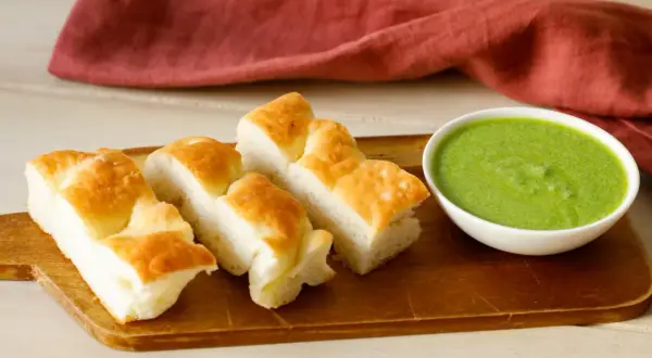 Focaccia tagliata a pezzi su tagliere di legno, accanto a una ciotola di pesto di salvia.