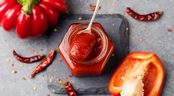 Un vasetto di marmellata di peperoni, con un cucchiaino che ne raccoglie un po'. Peperoni rossi interi e tagliati, peperoncini secchi e fiocchi di peperoncino completano la scena.