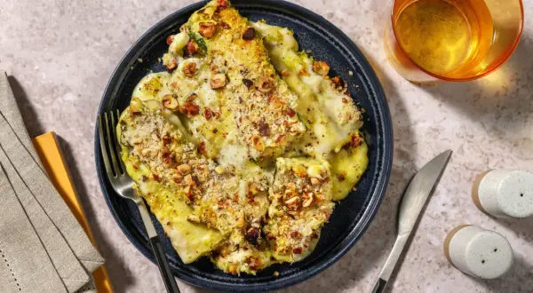 Parmigiana di zucchine gratinata con nocciole croccanti, servita su un piatto blu, con posate e un bicchiere di vino.