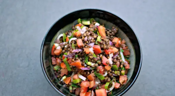 Una fresca insalata di lenticchie estiva, con pomodori, cipolla rossa e erbe aromatiche, servita in una ciotola scura.