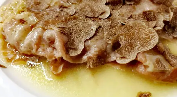 Cotoletta alla bolognese ricoperta di scaglie di tartufo, servita in un piatto bianco con una salsa dorata.