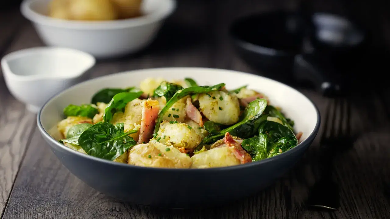 Insalata di patate con spinacini freschi e pancetta croccante, condita con erbe aromatiche. Un piatto gustoso e colorato.