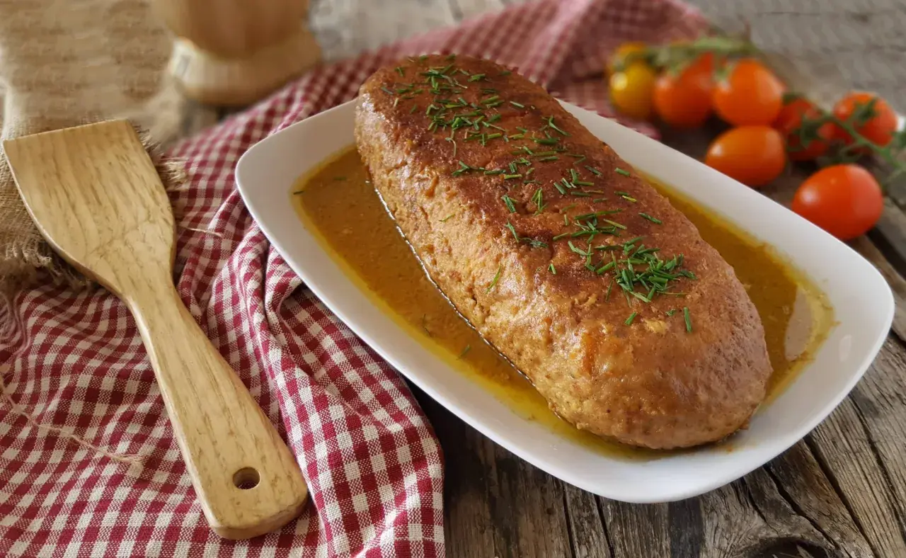 Polpettone in padella dorato e succulento, guarnito con erba cipollina, su un piatto bianco con salsa. Accanto, una spatola di legno e pomodorini.