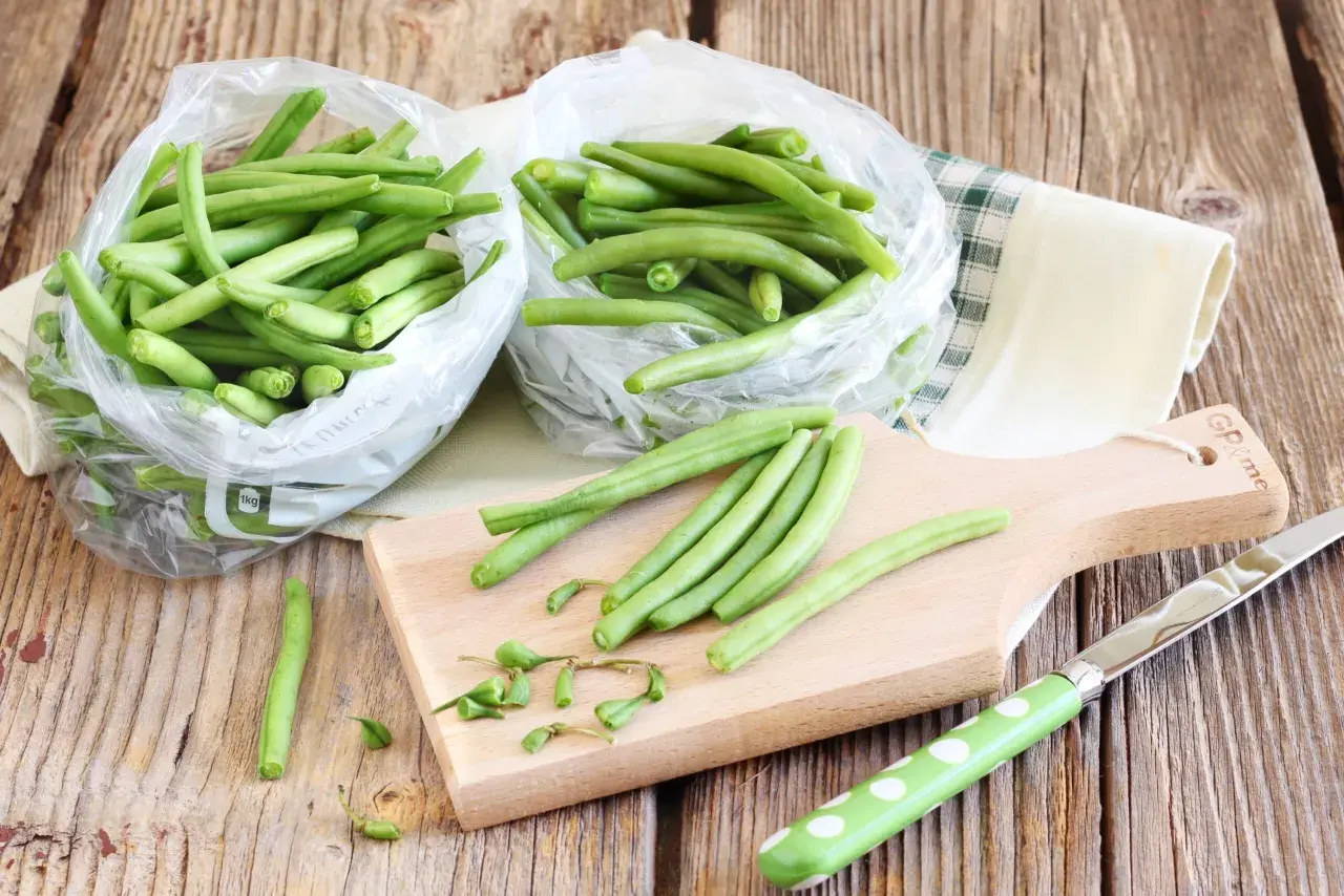 Fagiolini freschi in sacchetti trasparenti, pronti per essere preparati. Un tagliere di legno e un coltello suggeriscono come congelare i fagiolini per conservarli.
