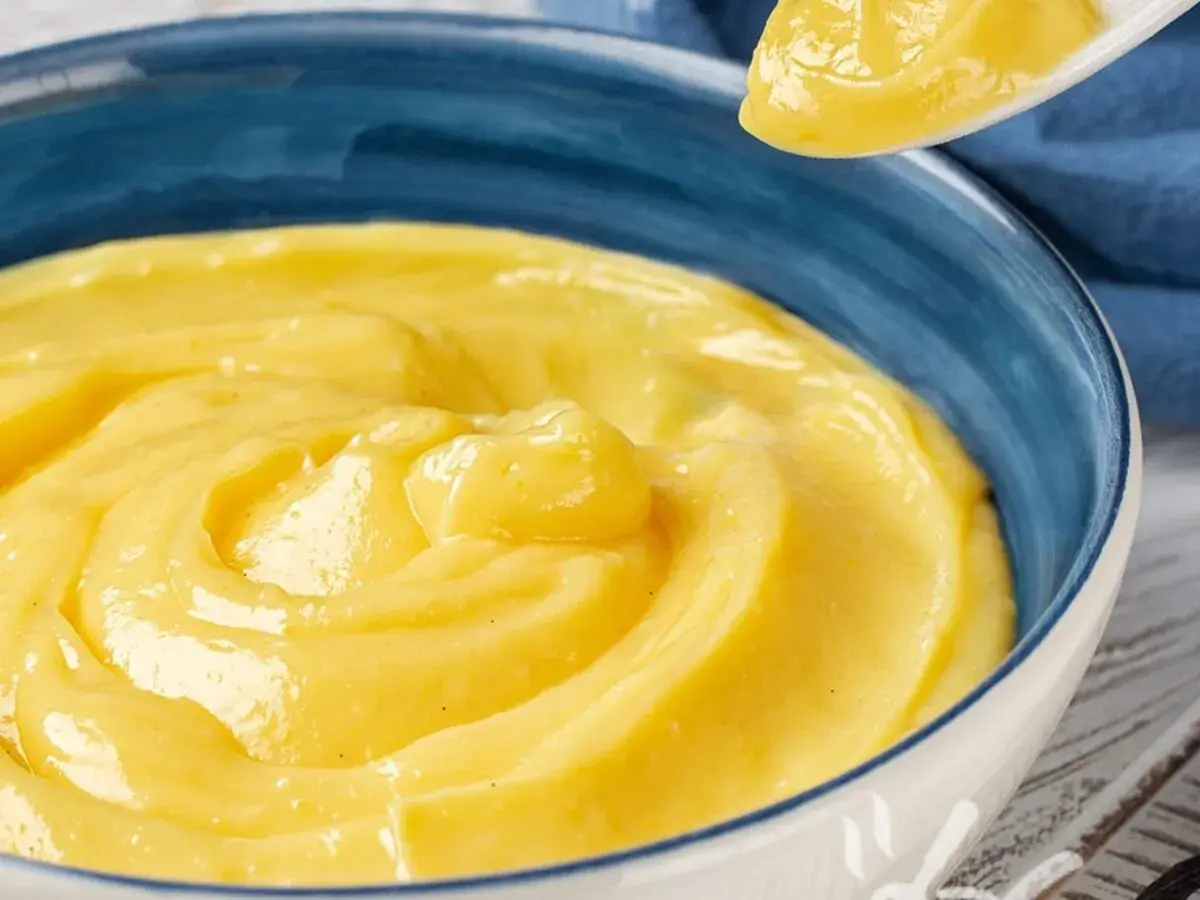 Una ciotola blu piena di vellutata crema pasticcera senza uova, con un cucchiaio che ne solleva una porzione.
