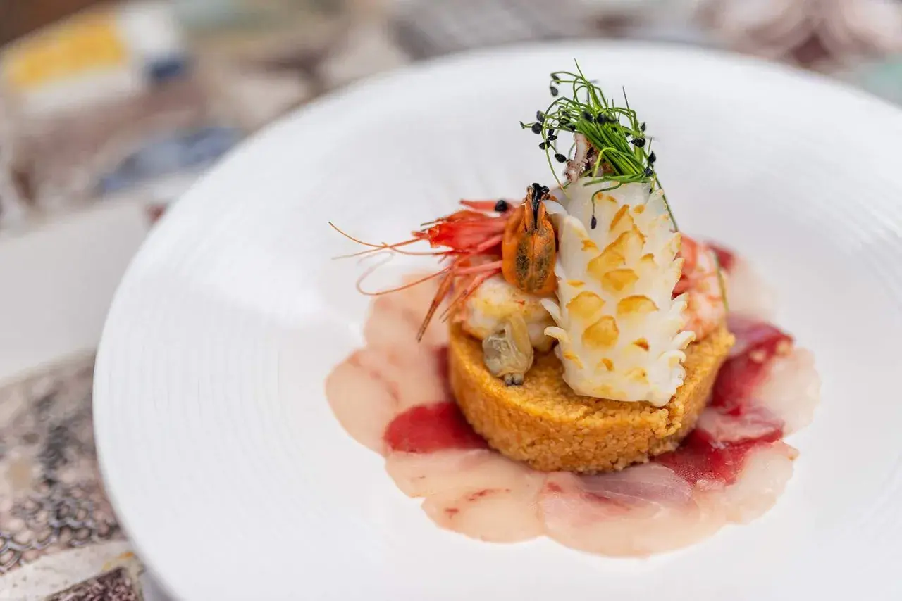 Un piatto elegante con una ricetta cous cous originale: gamberi, calamari e carpaccio di pesce su un letto di cous cous dorato, guarnito con erbe fresche.