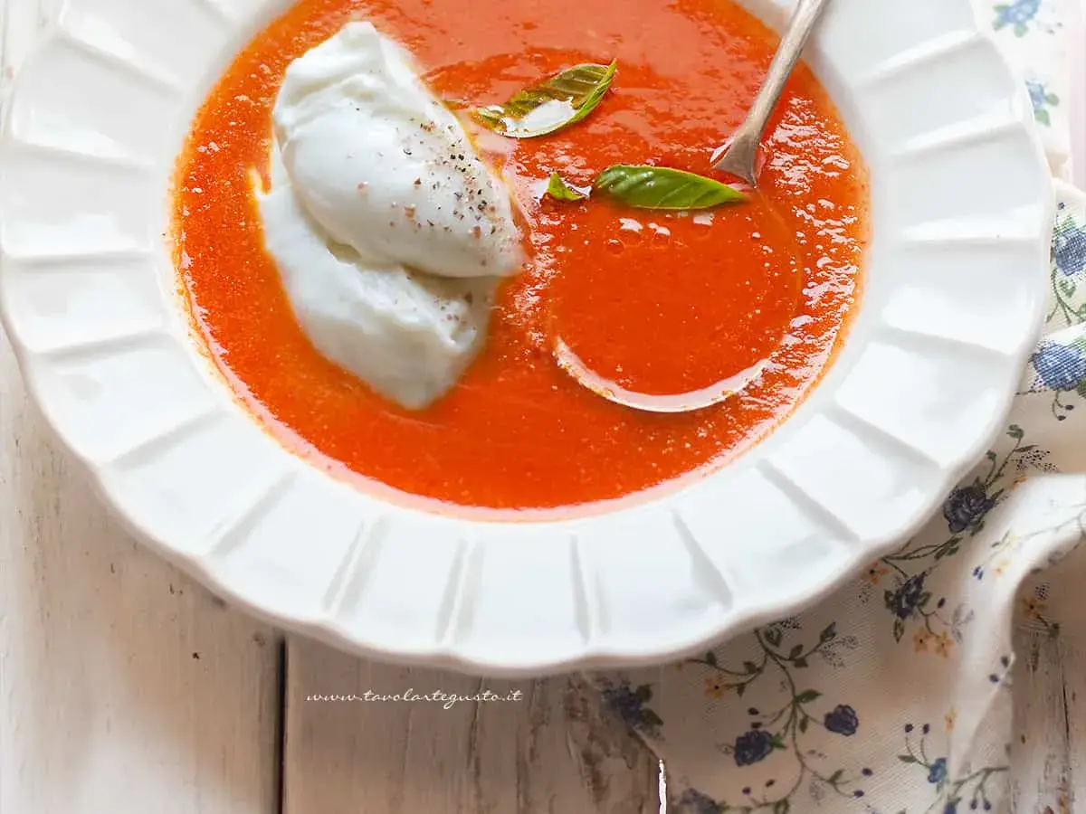 Una cremosa zuppa di pomodoro, arricchita da due quenelle di burrata e foglie di basilico fresco. Un piatto invitante e gustoso.
