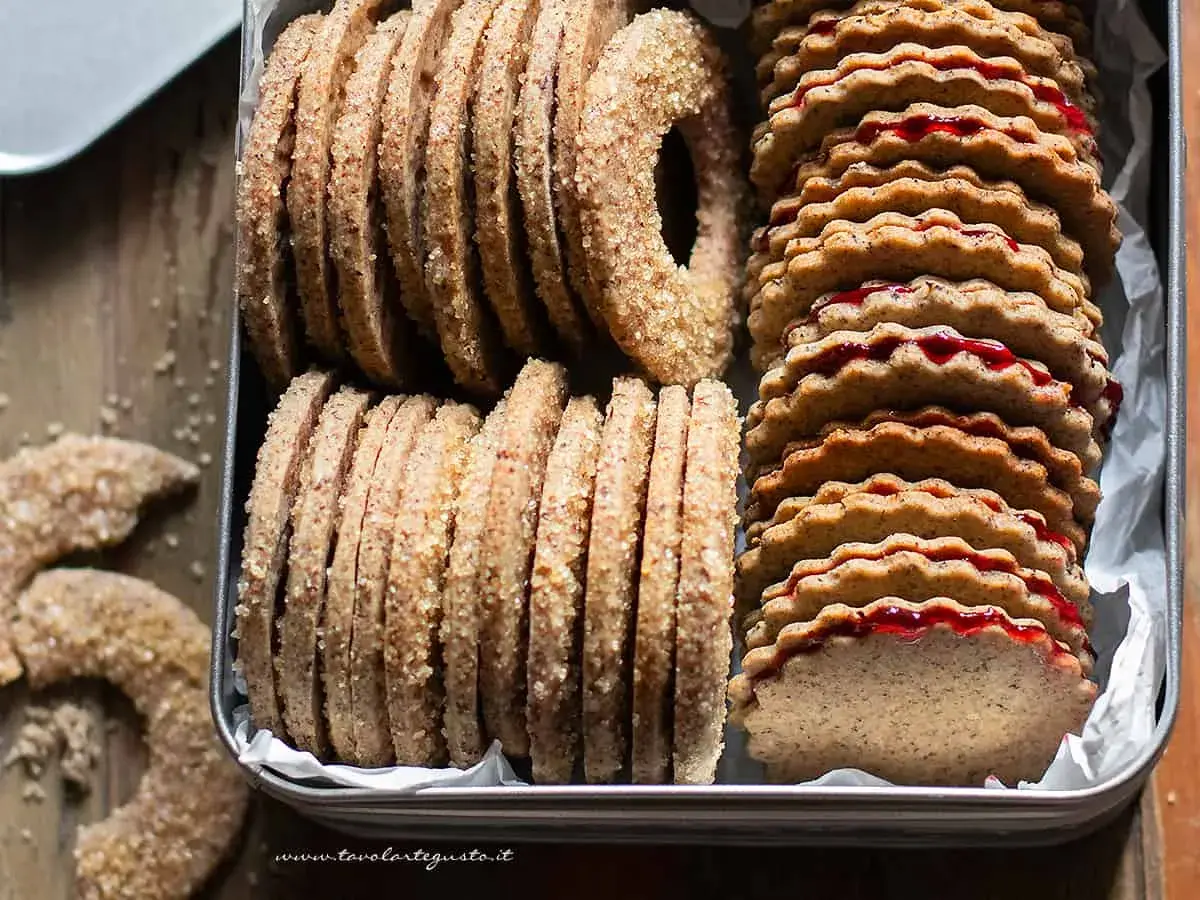 Biscotti grano saraceno ripieni di marmellata, impilati in una scatola di latta. Alcuni biscotti a forma di ciambella e a mezzaluna sono sparsi sul tavolo.