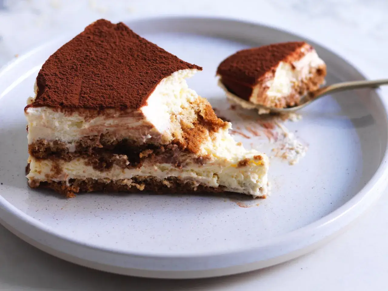 Una fetta di tiramisù ricetta originale, con strati di savoiardi, crema al mascarpone e cacao in polvere. Un cucchiaino ne ha già assaggiato un pezzetto.