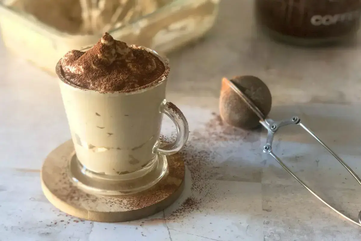 Deliziosa crema al caffè, spolverata di cacao, servita in una tazza di vetro con un colino per caffè accanto.