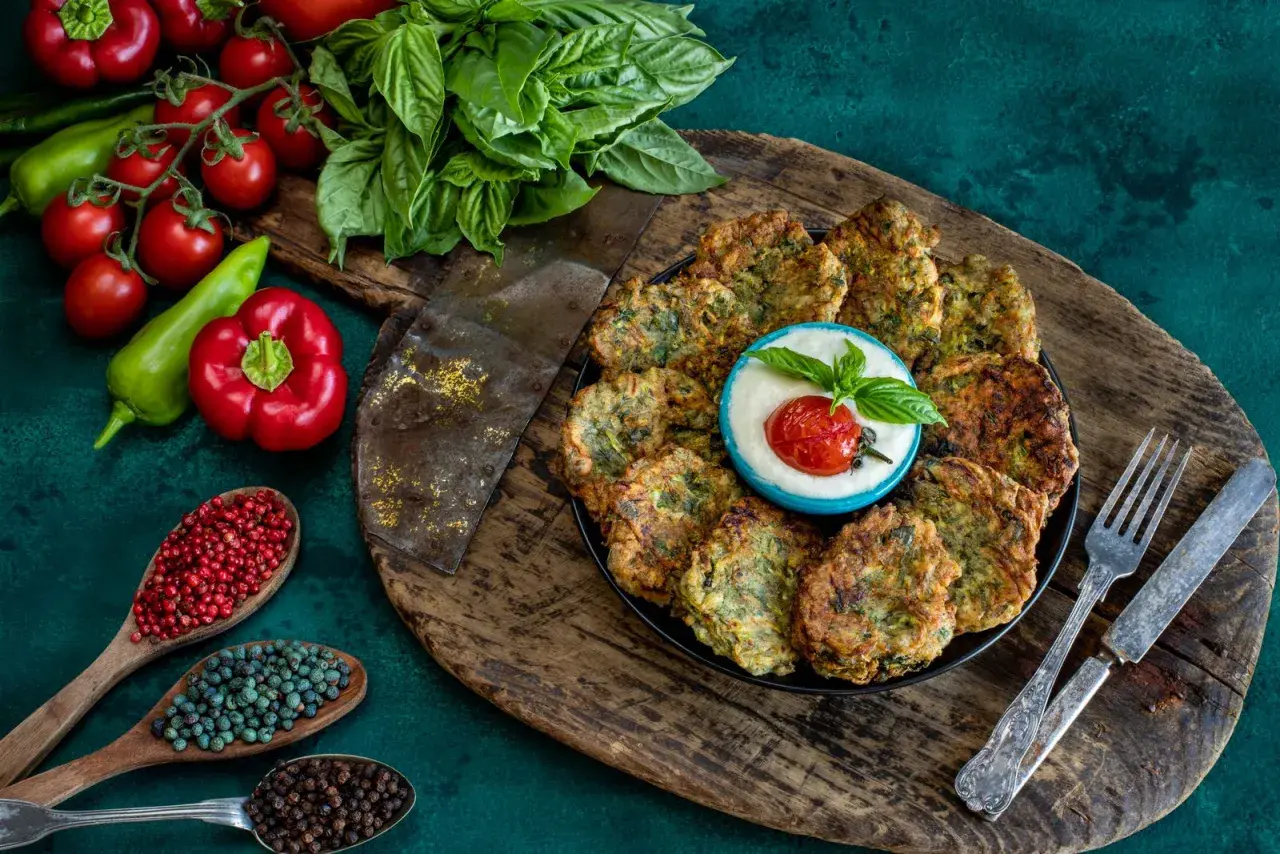 Piatto di polpette di zucchine dorate, servite con una ciotolina di salsa bianca, pomodorino e basilico. Ingredienti freschi e spezie completano la scena.