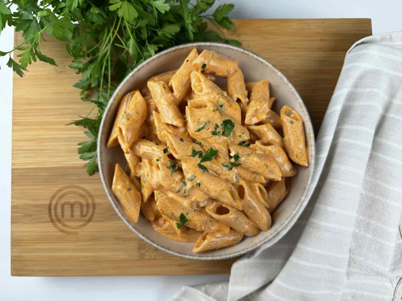 Penne alla vodka cremose e invitanti, guarnite con prezzemolo fresco, su un tagliere di legno con il logo di MasterChef.