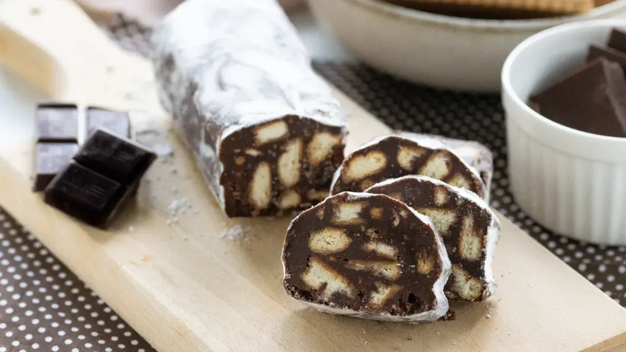 Un delizioso salame al cioccolato, cosparso di zucchero a velo, con biscotti e cioccolato fondente a lato.