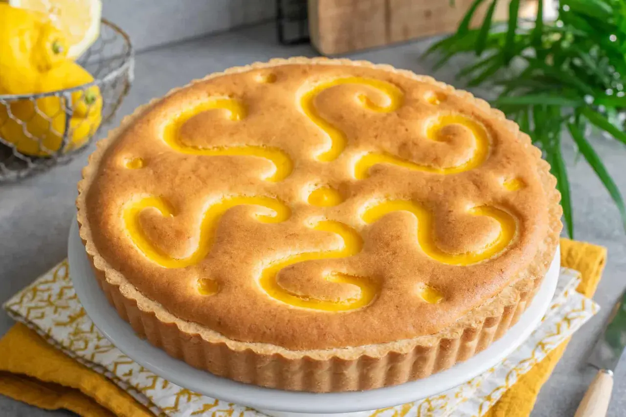 Torta con crema pasticcera, decorata con un motivo a spirale giallo brillante. Sullo sfondo, limoni freschi e foglie verdi.