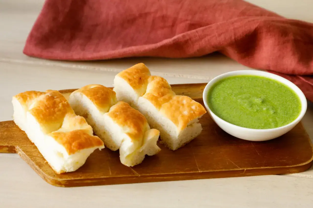Focaccia tagliata a pezzi su tagliere di legno, accanto a una ciotola di pesto di salvia.