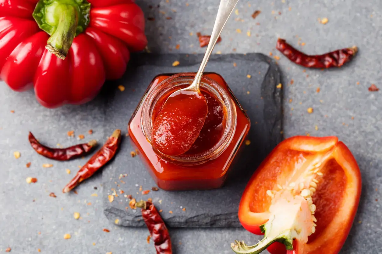 Un vasetto di marmellata di peperoni, con un cucchiaino che ne raccoglie un po'. Peperoni rossi interi e tagliati, peperoncini secchi e fiocchi di peperoncino completano la scena.