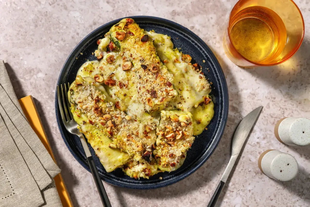 Parmigiana di zucchine gratinata con nocciole croccanti, servita su un piatto blu, con posate e un bicchiere di vino.
