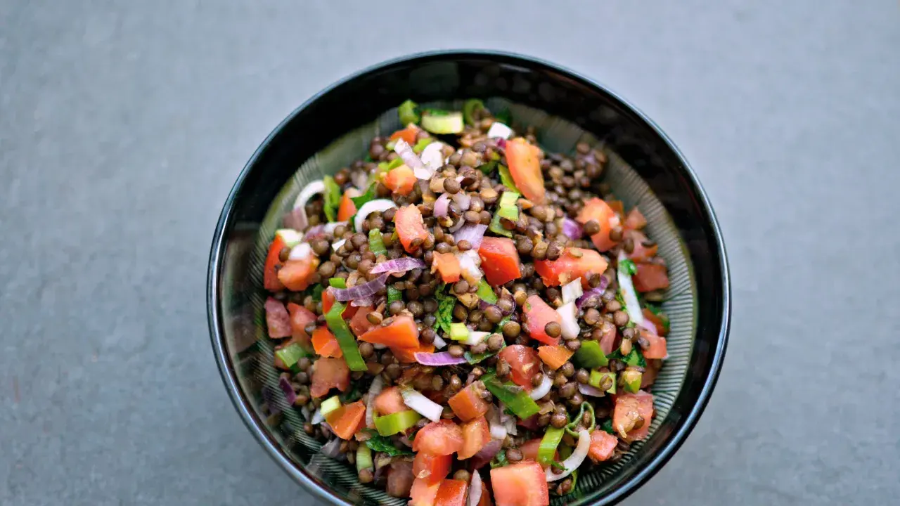 Una fresca insalata di lenticchie estiva, con pomodori, cipolla rossa e erbe aromatiche, servita in una ciotola scura.