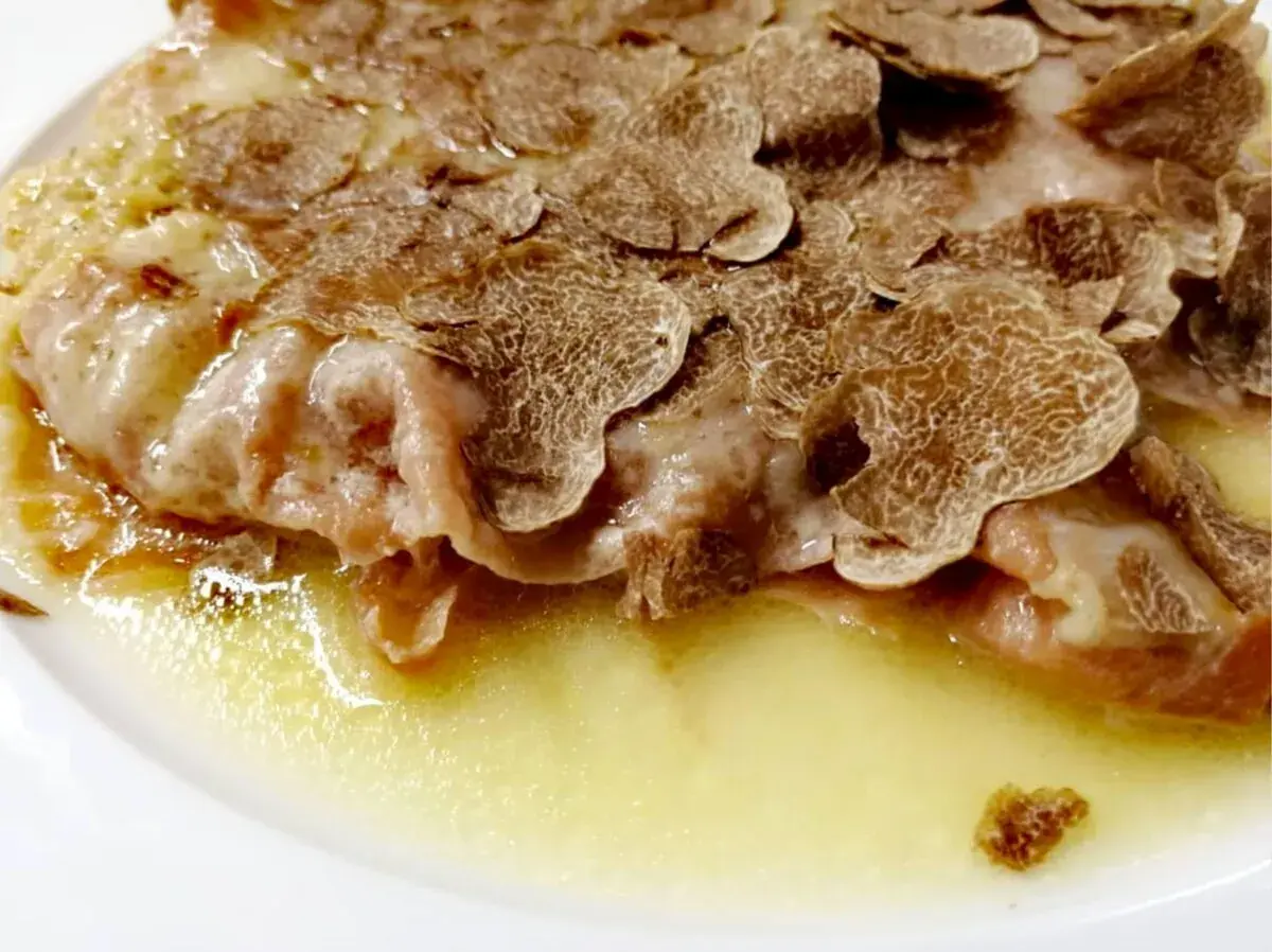 Cotoletta alla bolognese ricoperta di scaglie di tartufo, servita in un piatto bianco con una salsa dorata.