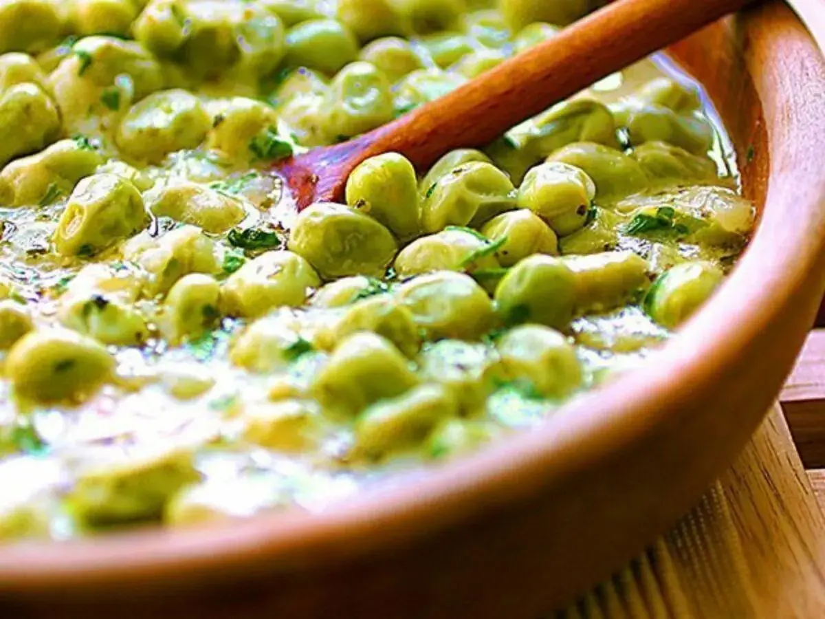 Un primo piatto saporito: piselli in umido, cremosi e profumati, serviti in una ciotola di legno con un cucchiaio.