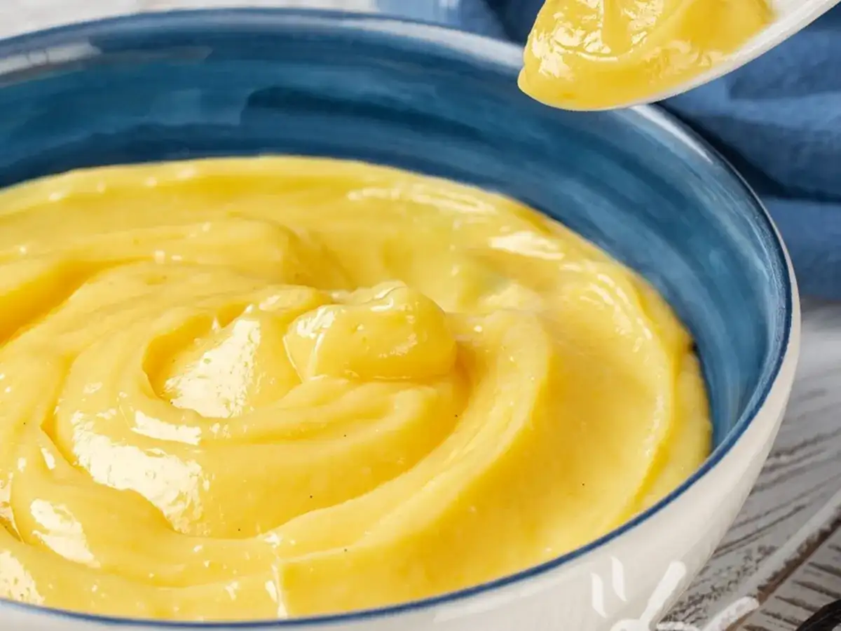 Una ciotola blu piena di vellutata crema pasticcera senza uova, con un cucchiaio che ne solleva una porzione.