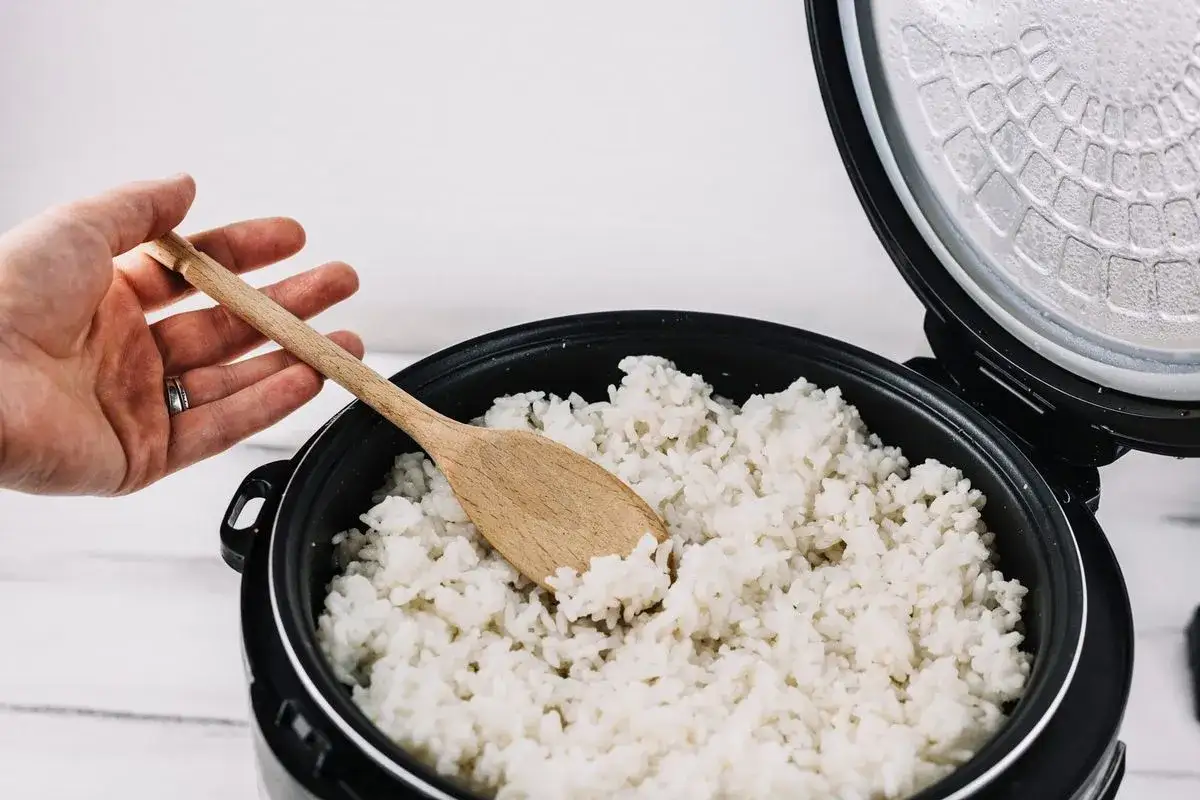Mano che mescola riso bianco cotto in una pentola con un cucchiaio di legno. Ecco come cuocere il riso perfettamente.