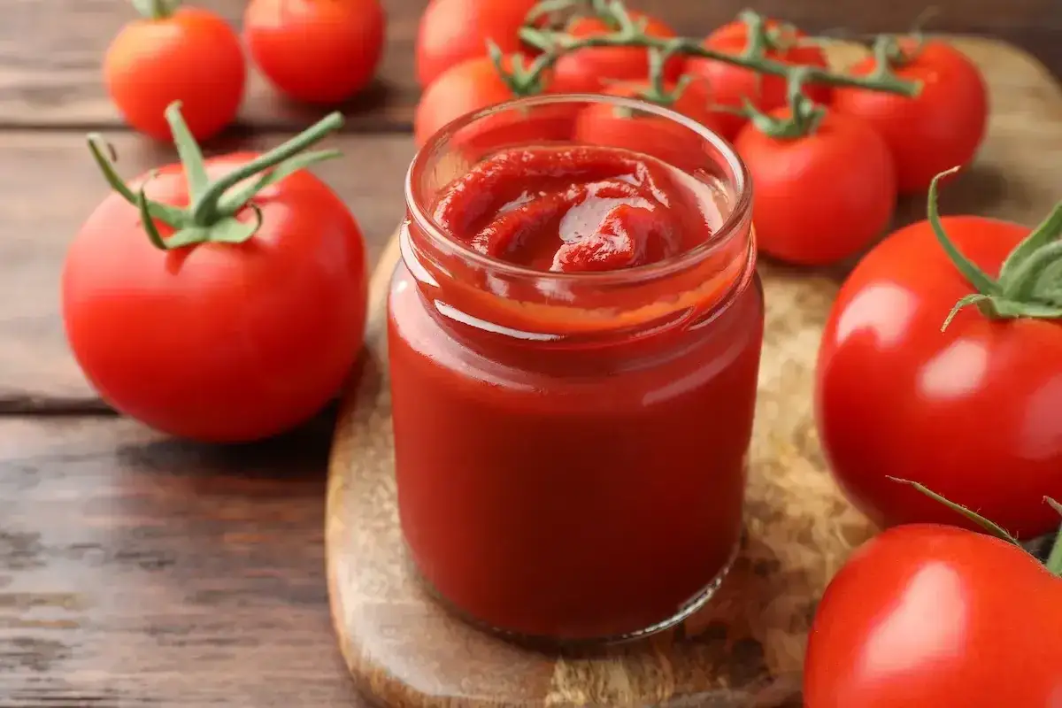 Un vasetto di ketchup fatto in casa, circondato da pomodori freschi su un tagliere di legno.