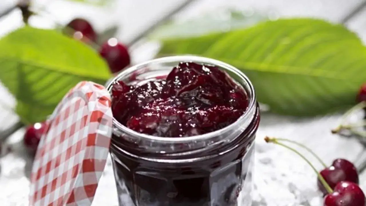 Vaso di marmellata di amarene, perfetta per la tua ricetta fatta in casa. Fresche amarene e foglie verdi sullo sfondo.