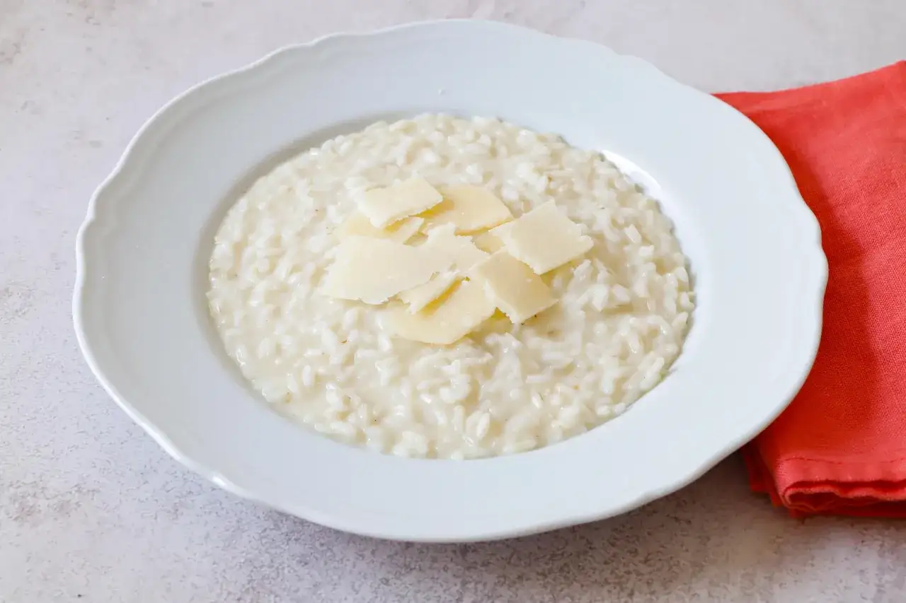 Un cremoso riso in bianco, guarnito con scaglie di parmigiano, servito su un piatto bianco con un tovagliolo arancione sullo sfondo.