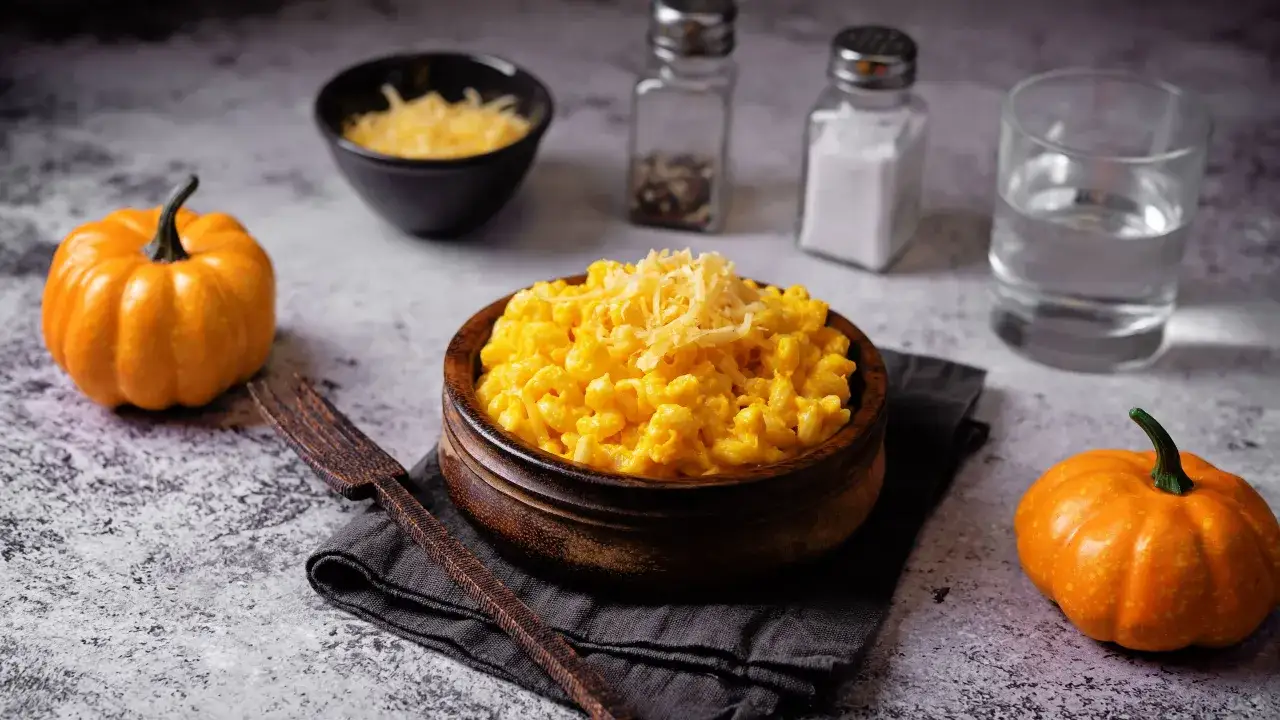 Un piatto di pasta e zucca cremoso, guarnito con formaggio grattugiato, accanto a piccole zucche arancioni.