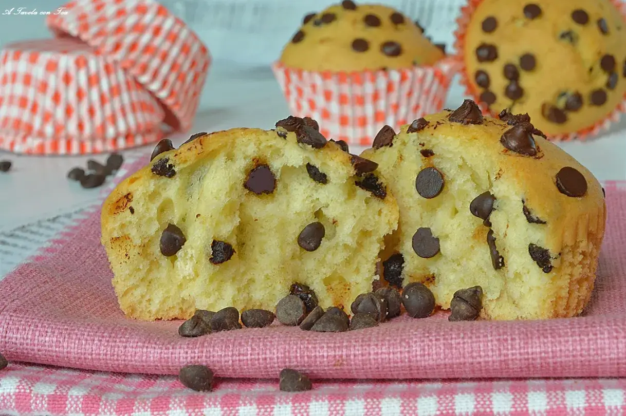 Muffin con gocce di cioccolato, uno tagliato a met&agrave;, su un tovagliolo rosa. Altri muffin e pirottini sullo sfondo.