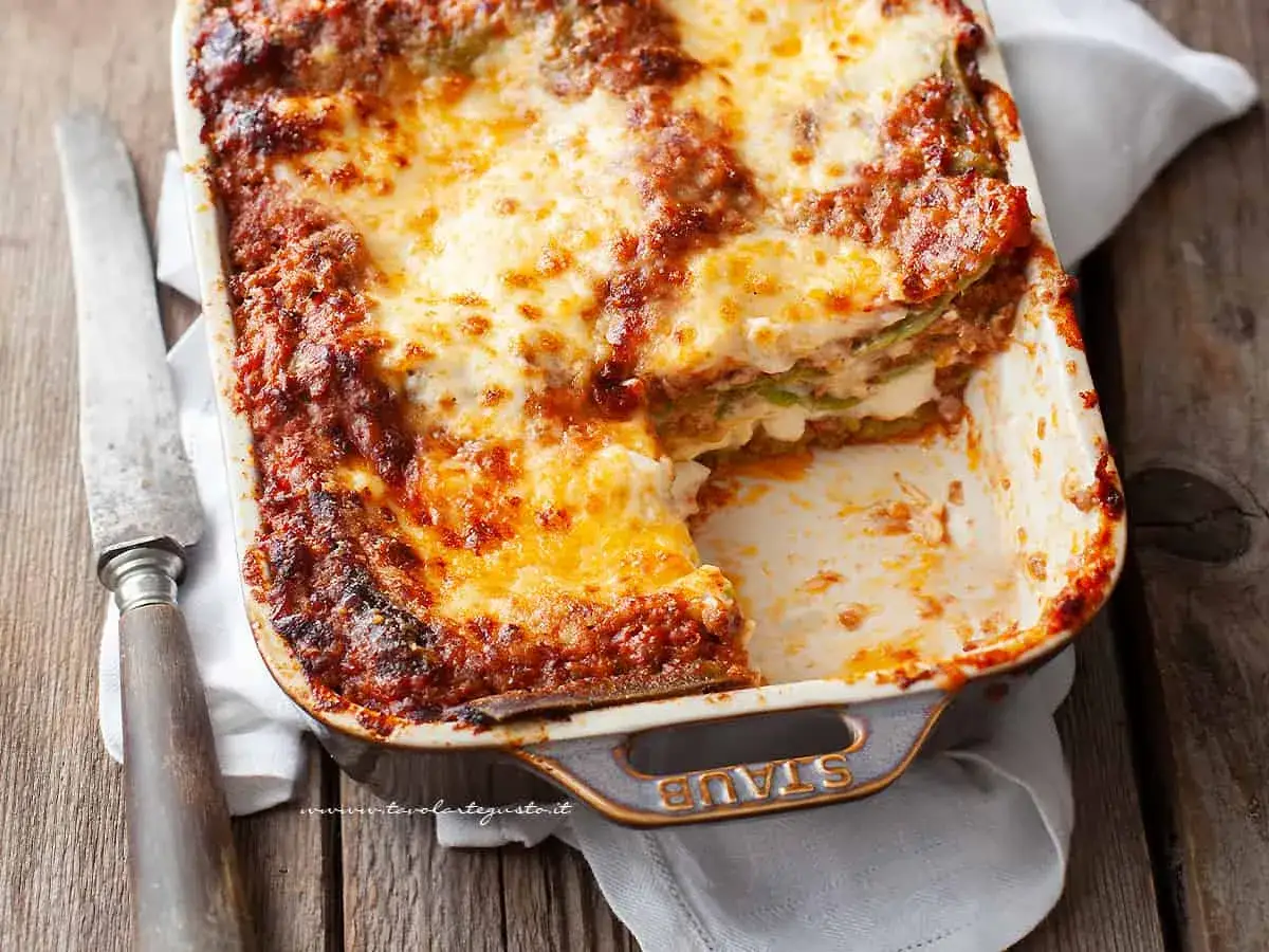 Lasagne alla bolognese appena sfornate, dorate e filanti, in una teglia Staub su un tavolo di legno.