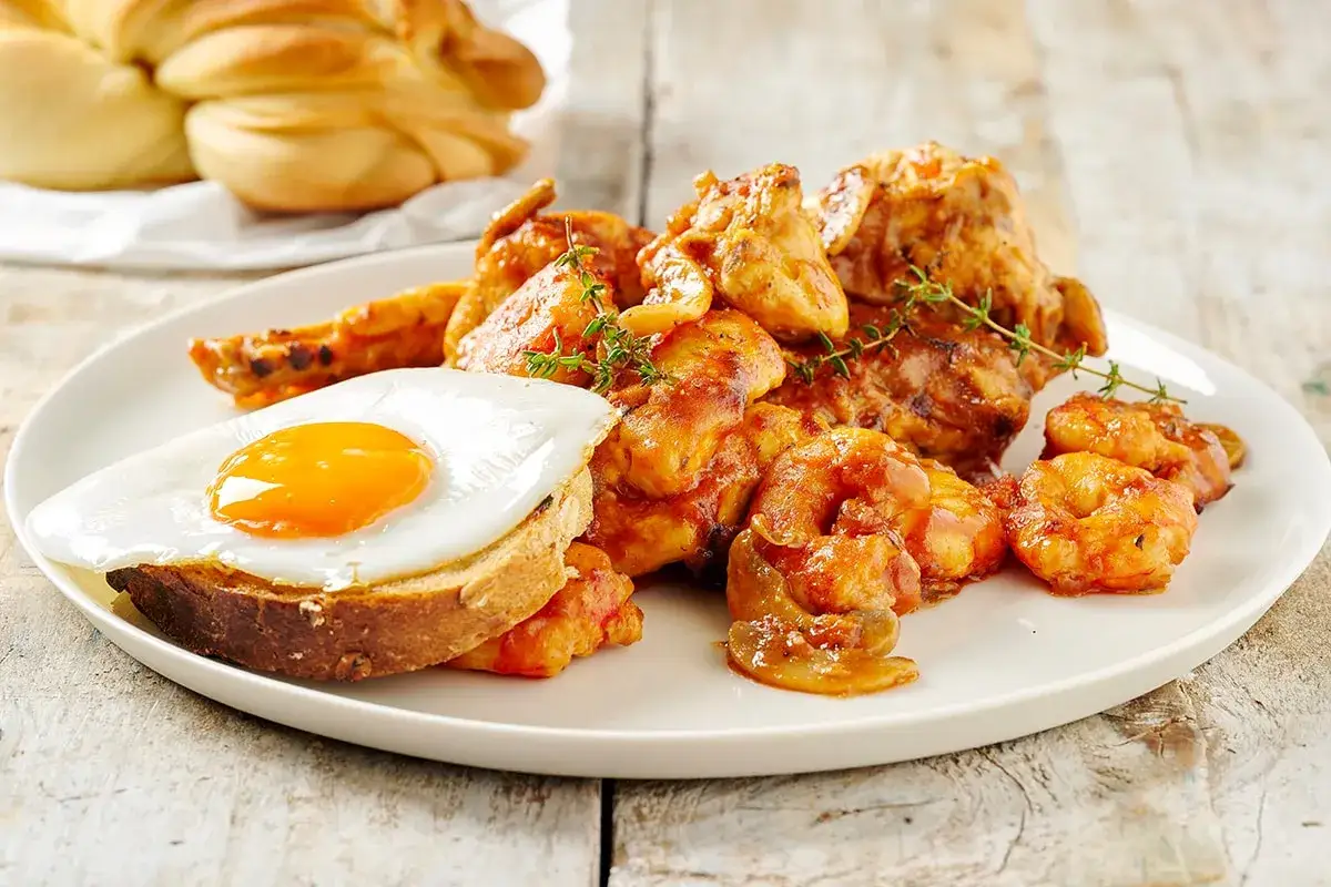 Piatto di pollo alla marengo con gamberi, uovo fritto su pane tostato e pane intrecciato sullo sfondo.