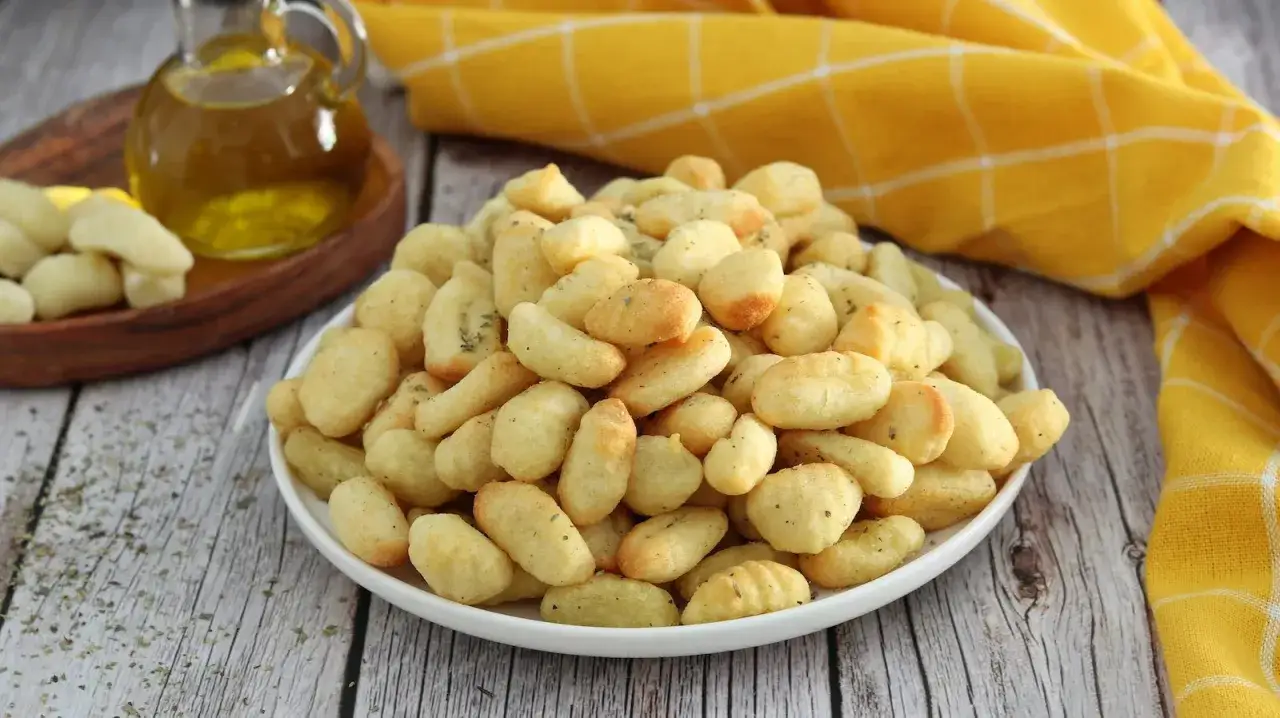 Gnocchi dorati e croccanti, preparati in friggitrice ad aria, conditi con erbe aromatiche. Accanto, un vasetto d'olio e gnocchi crudi.