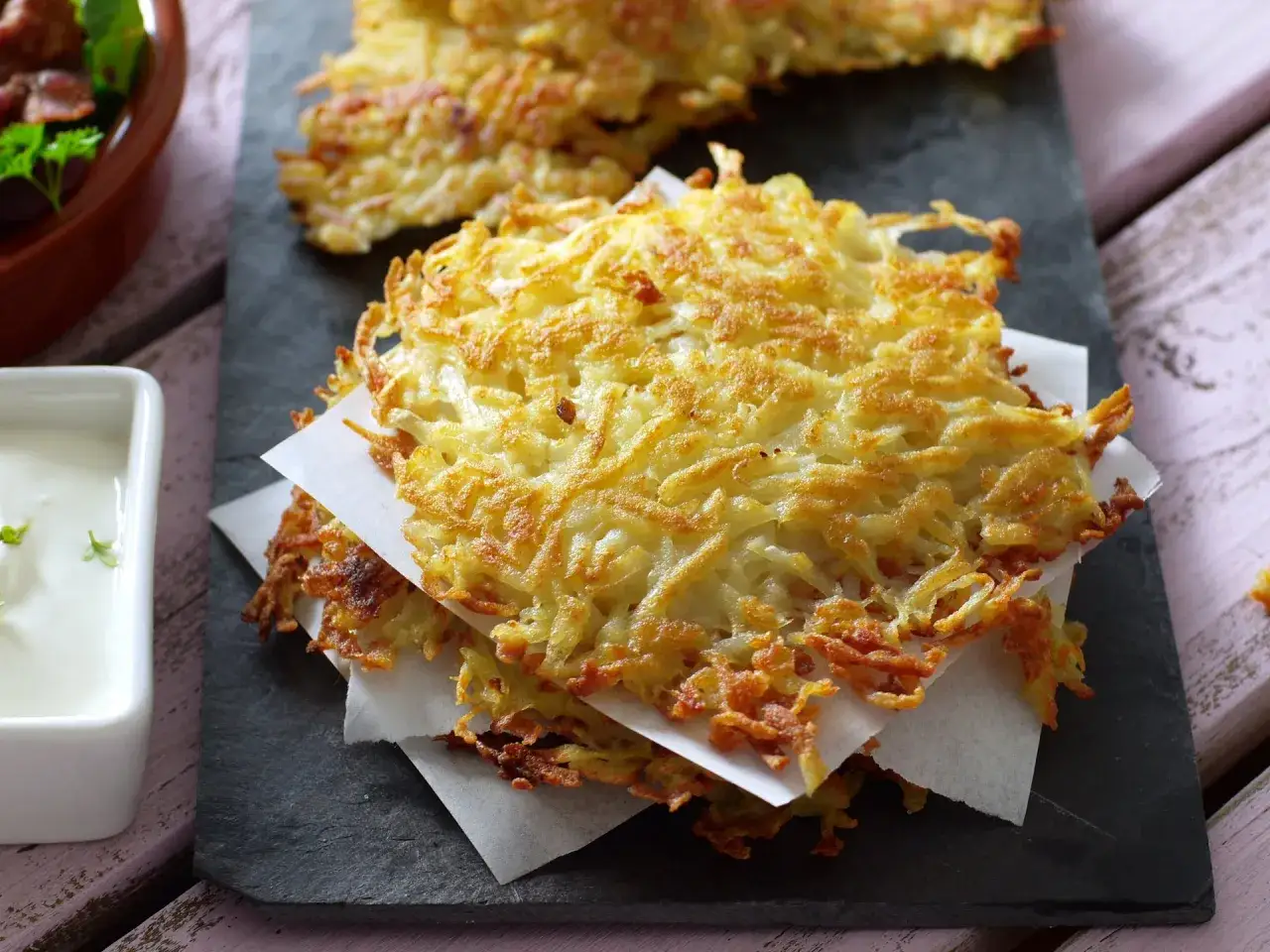 Deliziosi rosti di patate in padella, dorati e croccanti, serviti su una pietra scura con una ciotolina di salsa bianca.