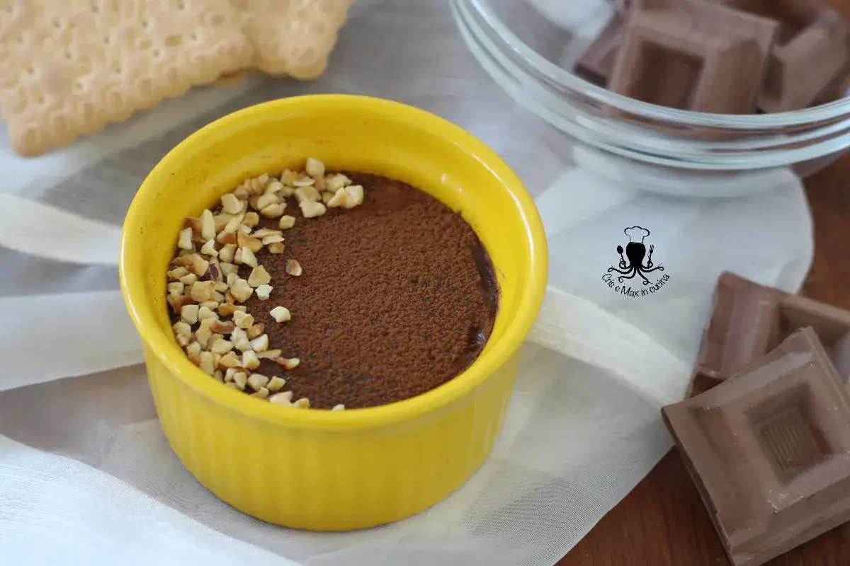 Budino al cioccolato in ciotola gialla, cosparso di cacao e nocciole tritate. Biscotti e cioccolato fondente sullo sfondo.
