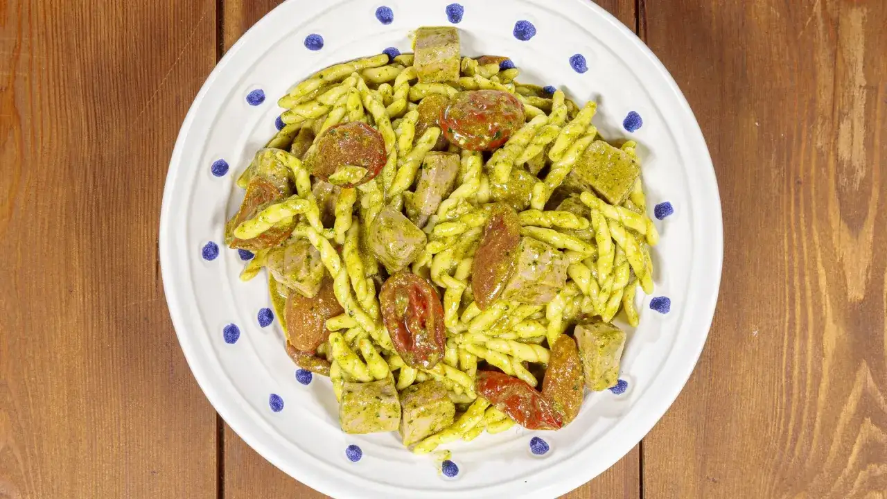 Piatto di trofie alla carlofortina con tonno, pomodorini secchi e pesto, servite su un piatto bianco decorato con pois blu.