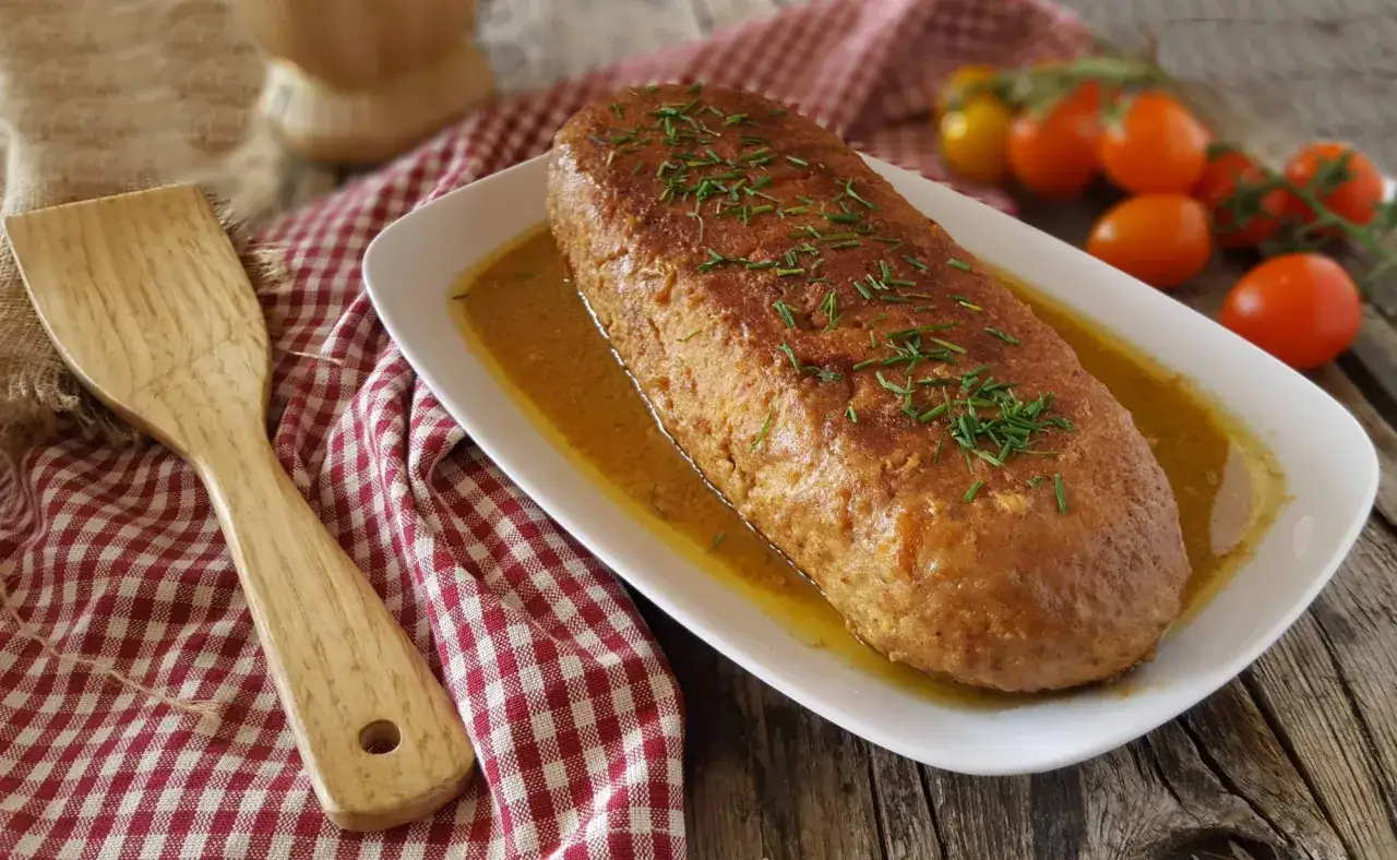 Polpettone in padella dorato e succulento, guarnito con erba cipollina, servito su un piatto bianco con un contorno di pomodorini.